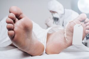 Feet of deceased person with blank ID tag on big toe, indicating coronavirus victim. Depressed doctor in protective gear mourns loss of patient.